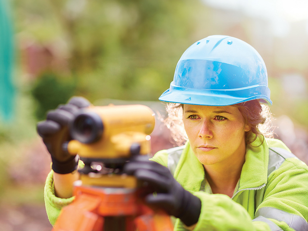 Woman wearing hard hat and high-vis jacket looking through levelling telescope